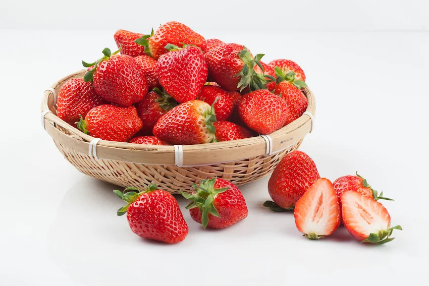 하얀 배경 위 나무 바구니에 가득 담긴 신선한 빨간 딸기들과 바닥에 놓인 딸기 단면 조각