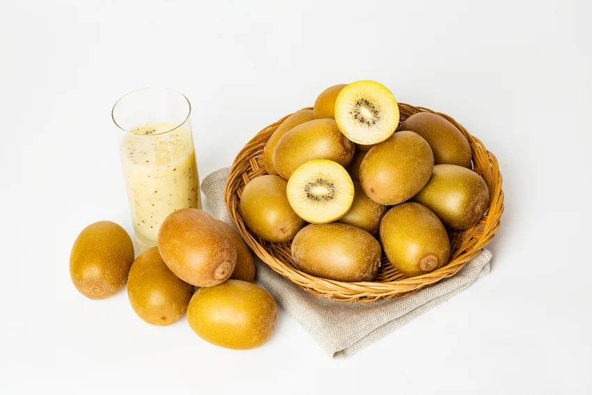 흰색 배경 위 대바구니에 담긴 여러 개의 골드키위와 반으로 잘린 단면, 옆에 놓인 신선한 키위 생과일주스 한 잔