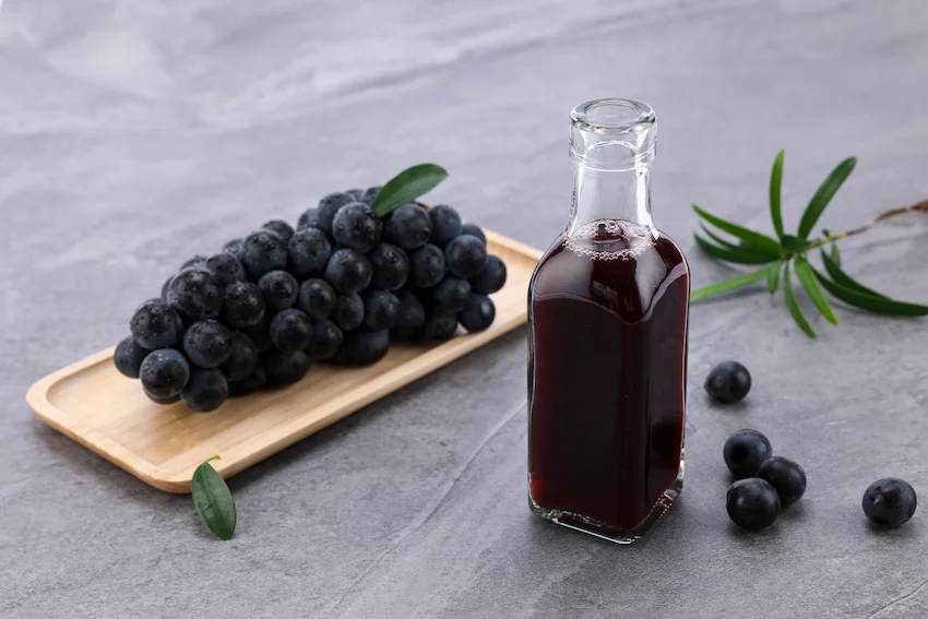 회색 석판 위 나무 쟁반에 담긴 검은 포도 한 송이와 유리병에 담긴 진한 보라색 포도 주스