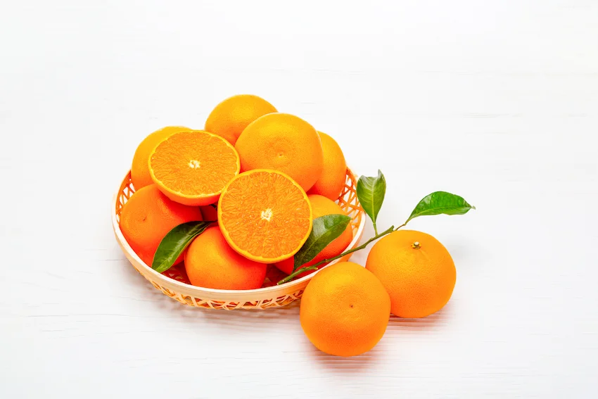 흰색 배경 위 대바구니에 가득 담긴 주황빛 천혜향과 반으로 잘린 과육 단면 및 초록색 잎사귀가 달린 가지