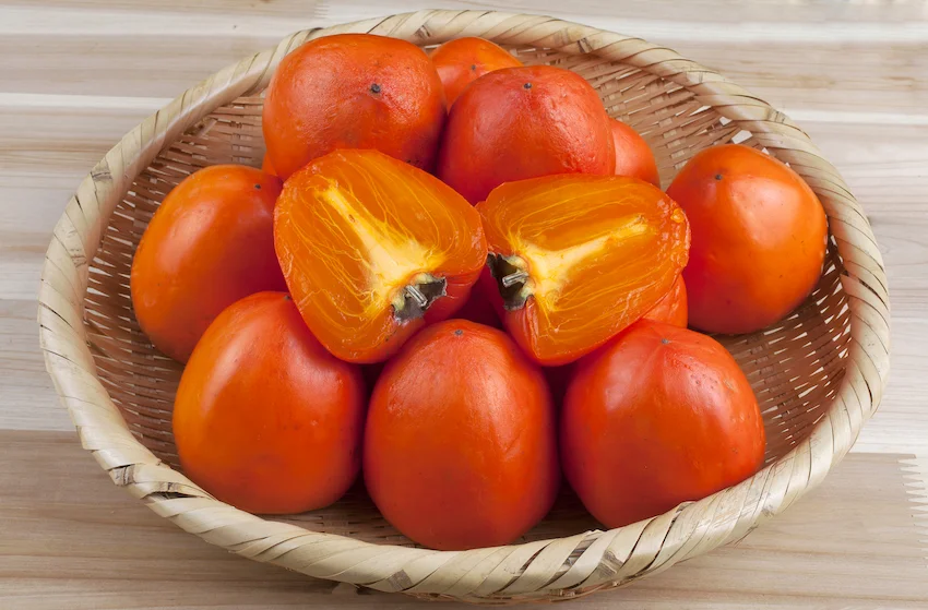 나무 테이블 위 대바구니에 가득 담긴 잘 익은 붉은 홍시들과 반으로 갈라진 대봉감의 촉촉한 속살