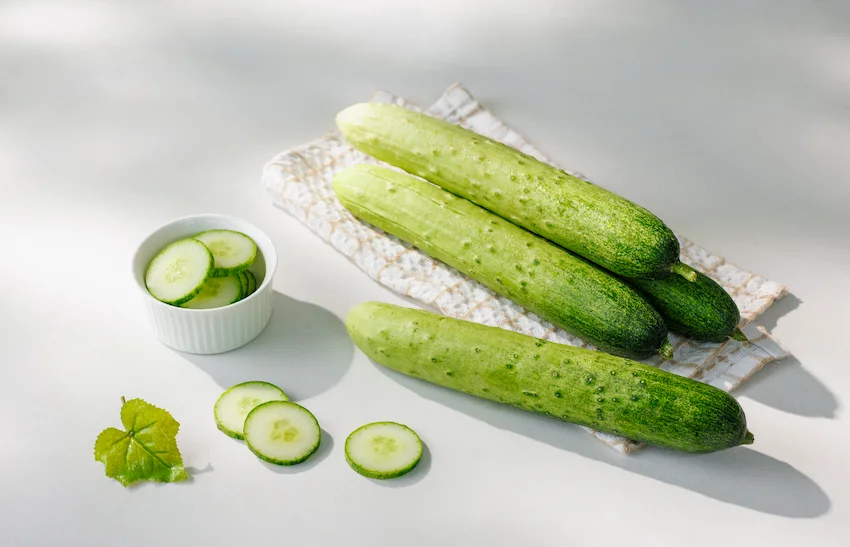 오이, 밝고 깨끗한 배경 위에 천에 올라간 싱싱한 통오이들과 작은 그릇에 담긴 동글동글하게 썰린 오이 슬라이스가 깔끔하게 놓여 있는 연출 컷 사진